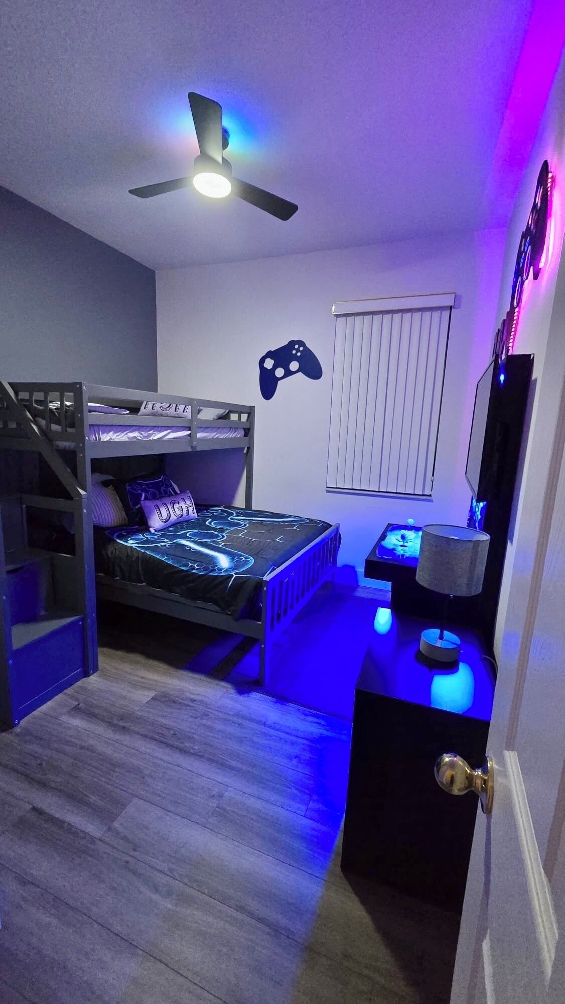 Une chambre avec des lits superposés, un ventilateur de plafond, une console de jeux et une fenêtre avec des stores.