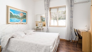 Quarto com cama, escrivaninha, cadeira, janela com cortinas e um quadro na parede.