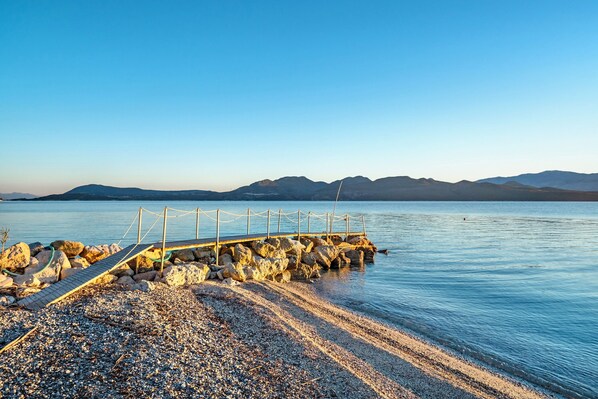 A pebbled beach with a wooden pier extending into calm water, surrounded by mountains.