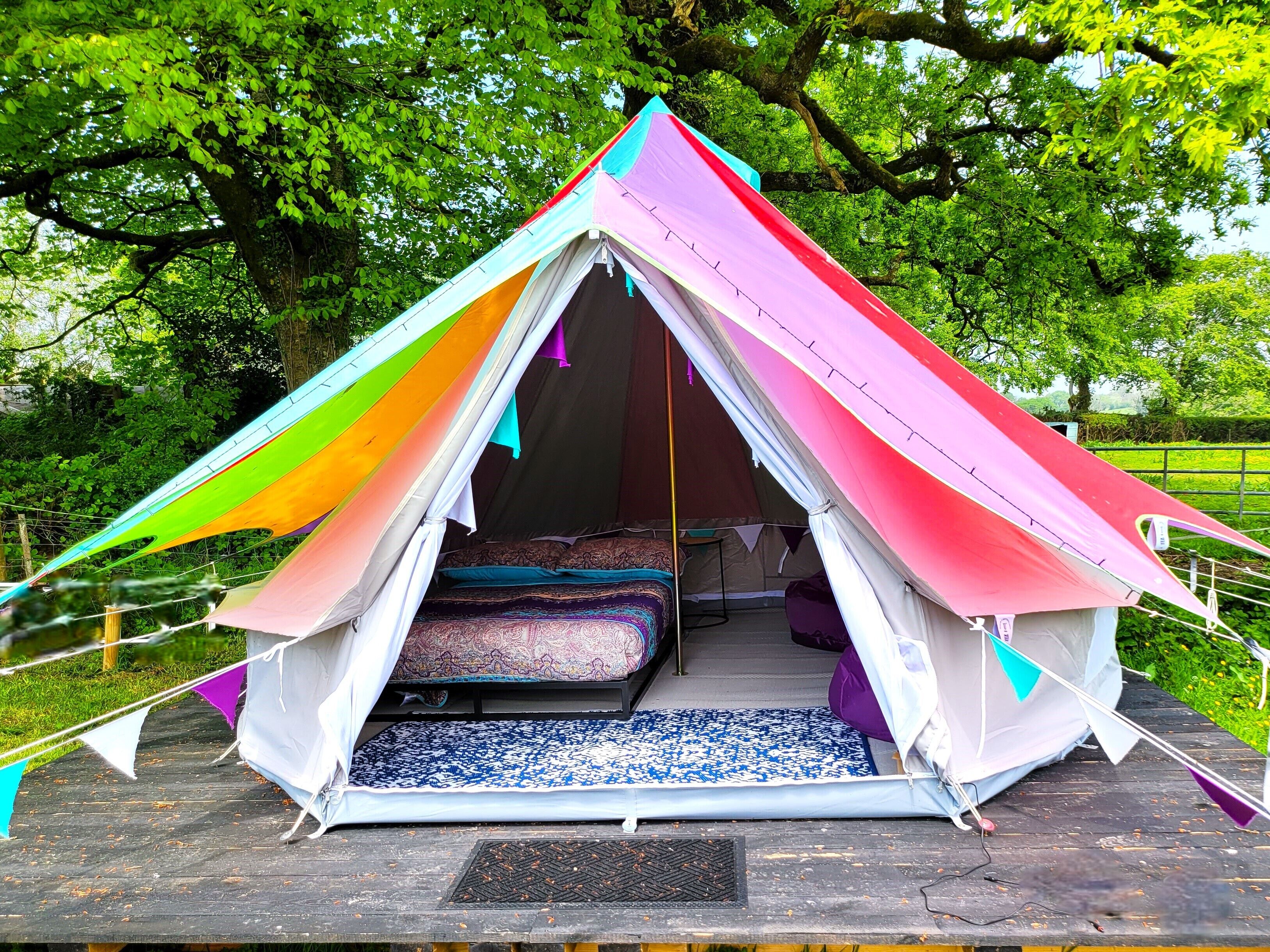 A colorful tent with a bed inside, set on a wooden platform in a green area.