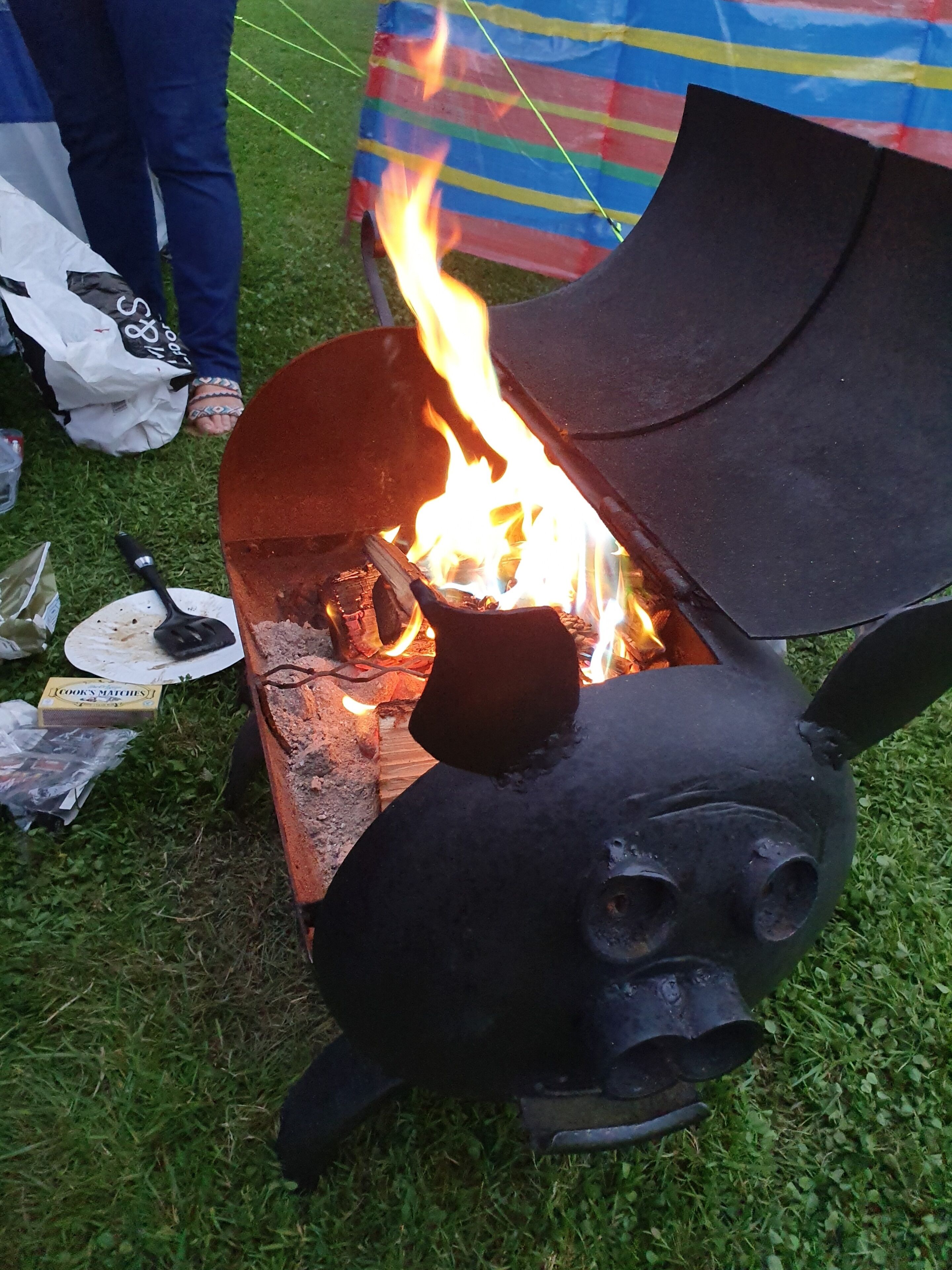Un brasero metálico con diseño facial, leña ardiendo y un fondo de tela a rayas.