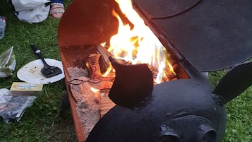 Un brasero metálico con diseño facial, leña ardiendo y un fondo de tela a rayas.