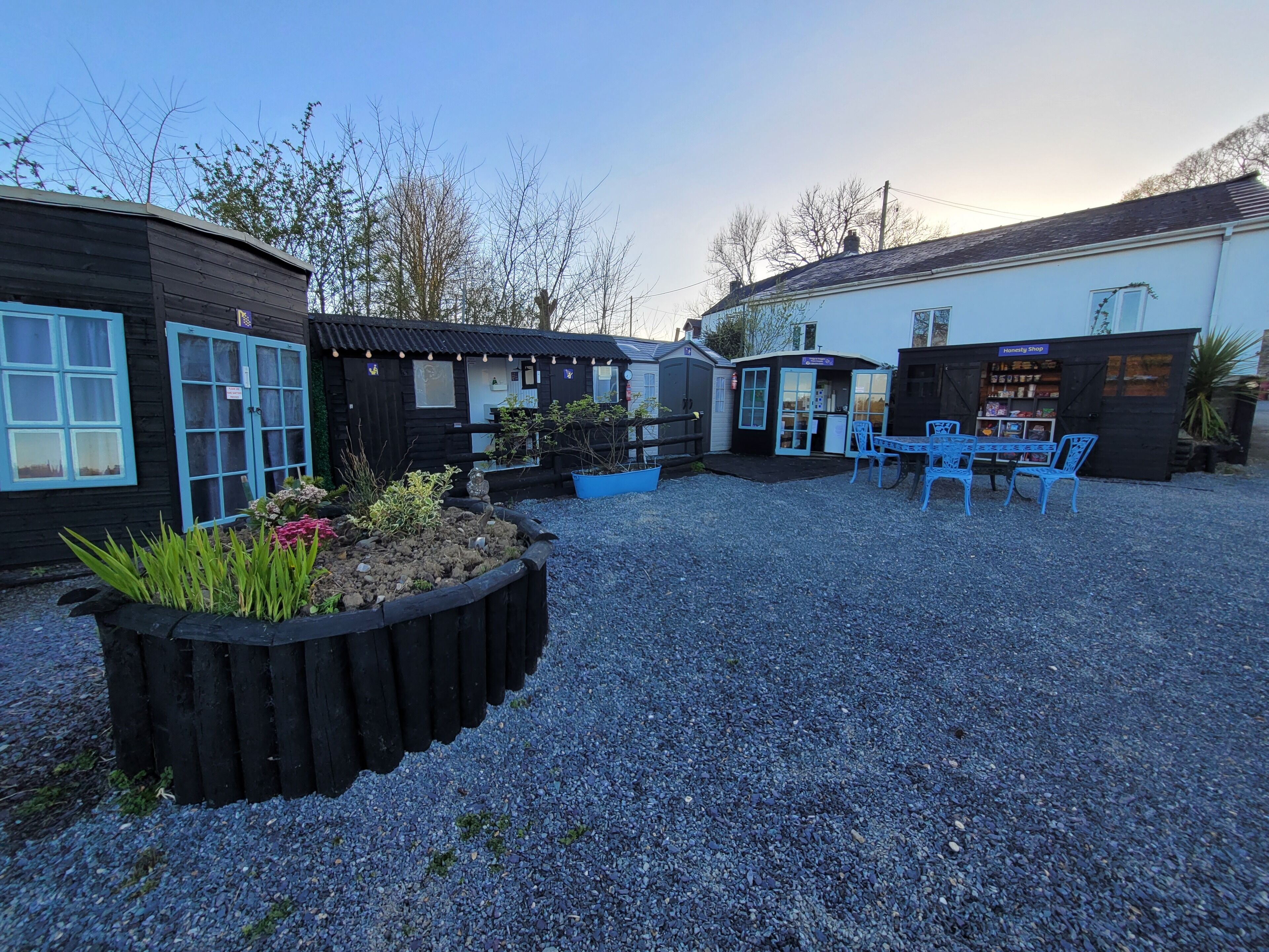 A courtyard with a gravel ground, wooden buildings, and a seating area with blue chairs.