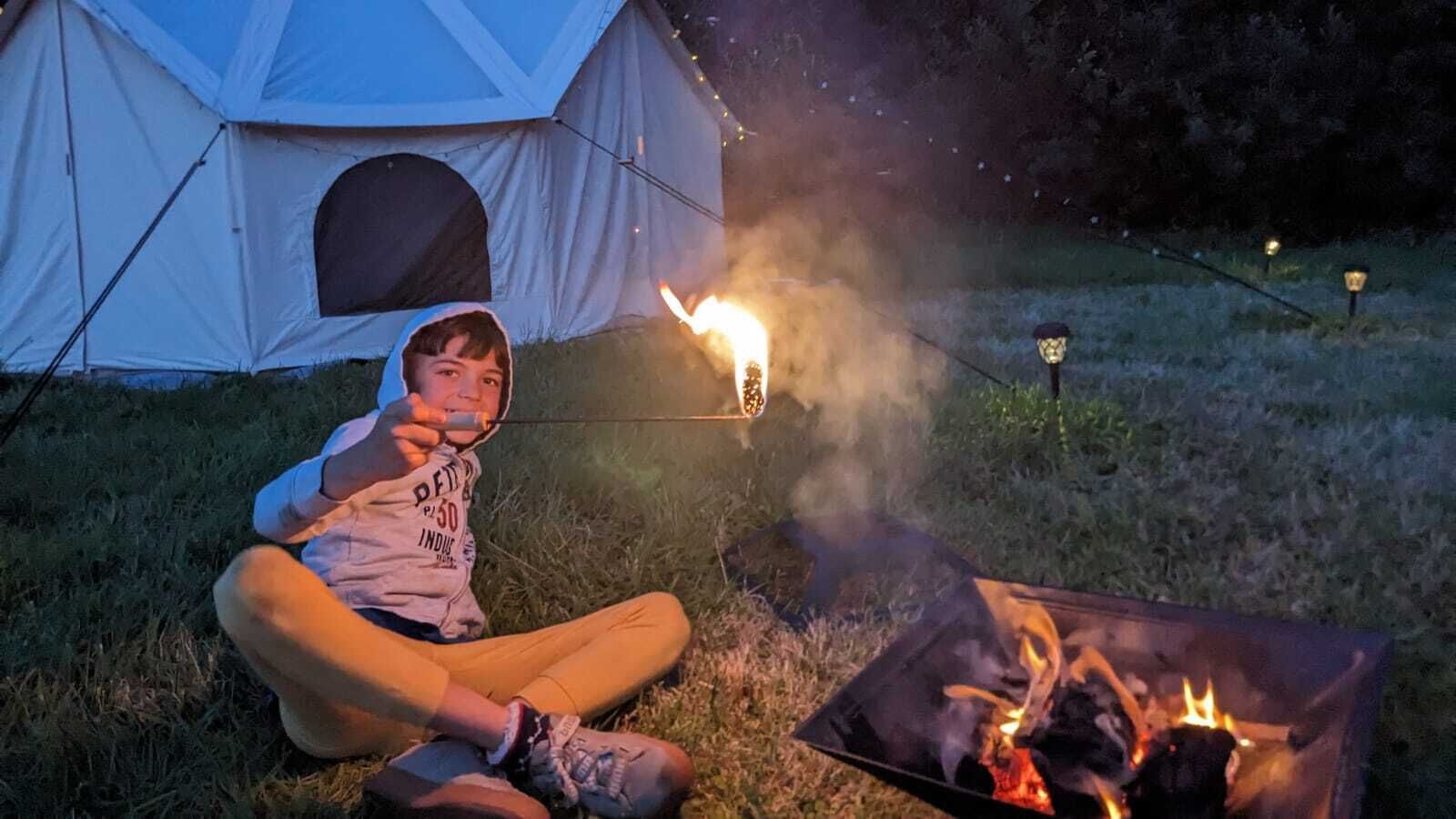 A person sitting by a campfire with a tent and a portable stove in the background.
