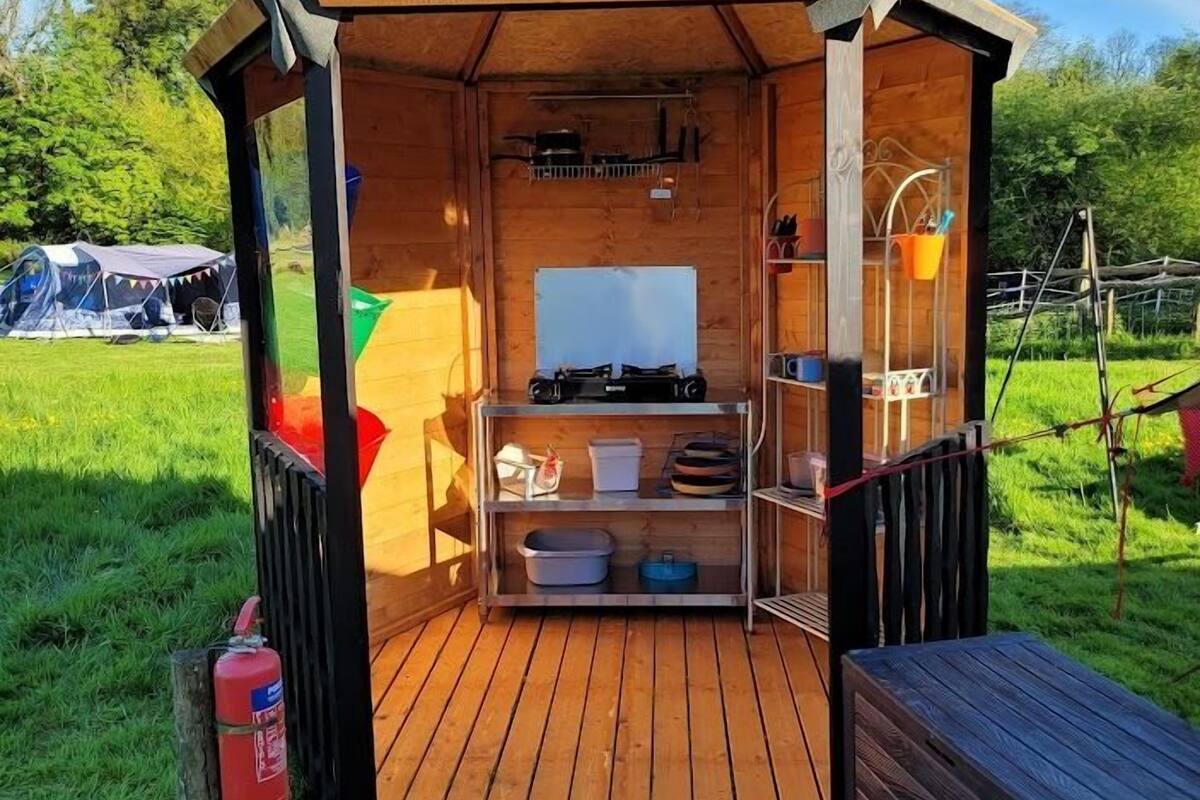 Een klein houten bouwwerk met een raam met metalen frame, een plank met keukengerei, en een brandblusser erbuiten.