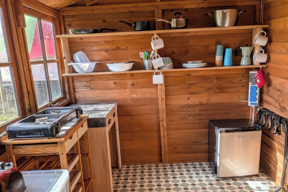 Een kleine keuken met houten wanden, een betegelde vloer en voorzieningen zoals een kookplaat, koelkast en planken met keukengerei.