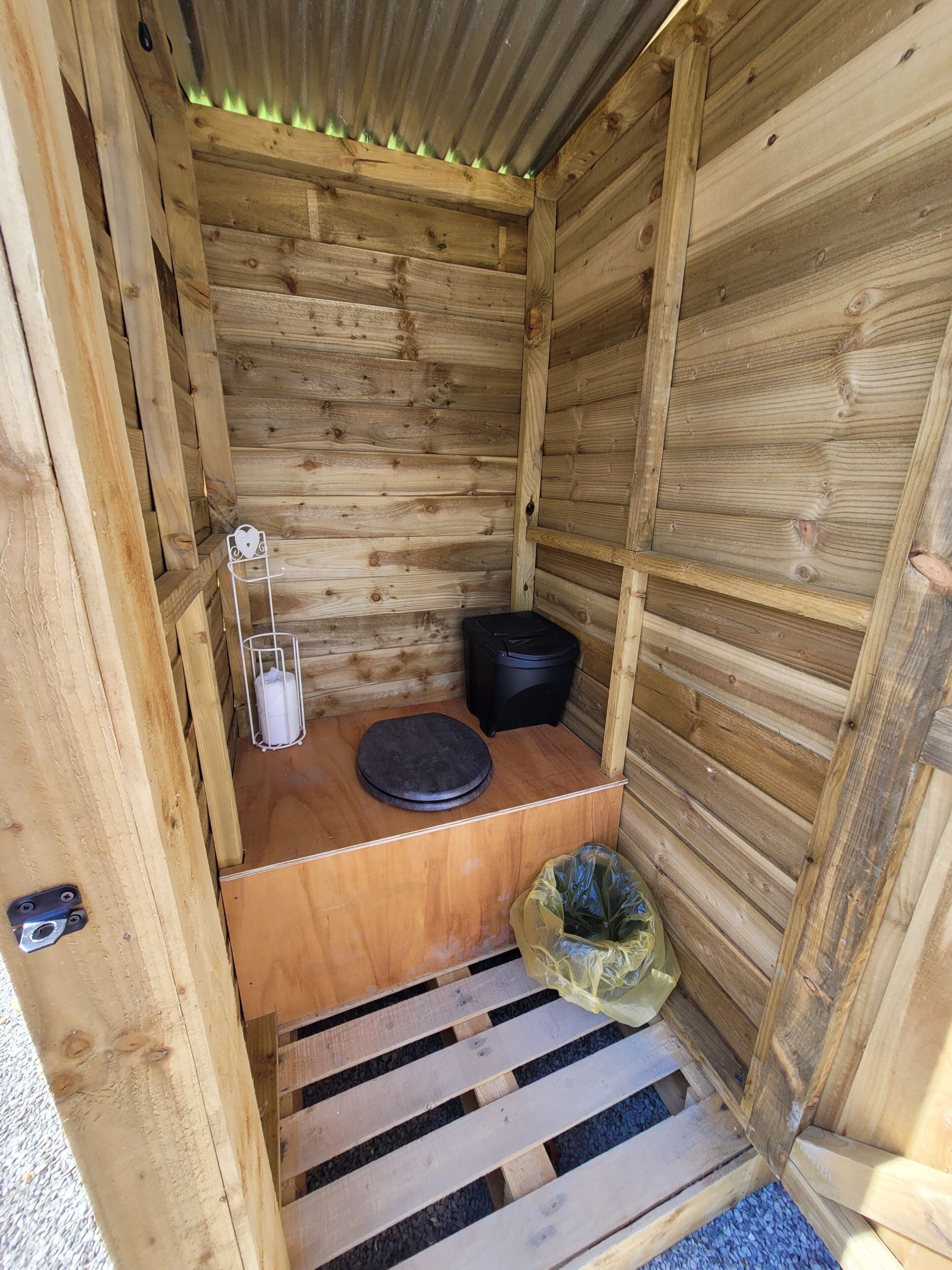 Una cabaña de madera con un banco, un bote de basura negro y un bote de basura blanco.