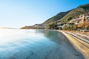 A coastal road with a pebble beach, houses on a hillside, and a clear sky. - Studio Apartment 'Old Boat Double Studio 8' with Sea View, Wi-Fi and Air Conditioning (Lefkada)