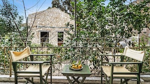 Ein Balkon mit zwei grünen Stühlen und einem kleinen Tisch, im Hintergrund ein Steinhaus und ein Metallzaun.