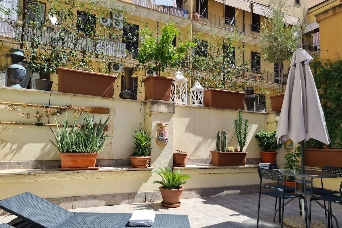 Un patio al aire libre con varias macetas, una mesa con sillas y una sombrilla.