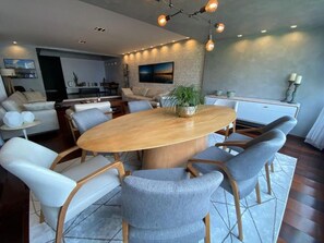 Una sala moderna con una mesa redonda de madera rodeada por seis sillas grises tapizadas, un sofá y una zona de comedor con un armario blanco.