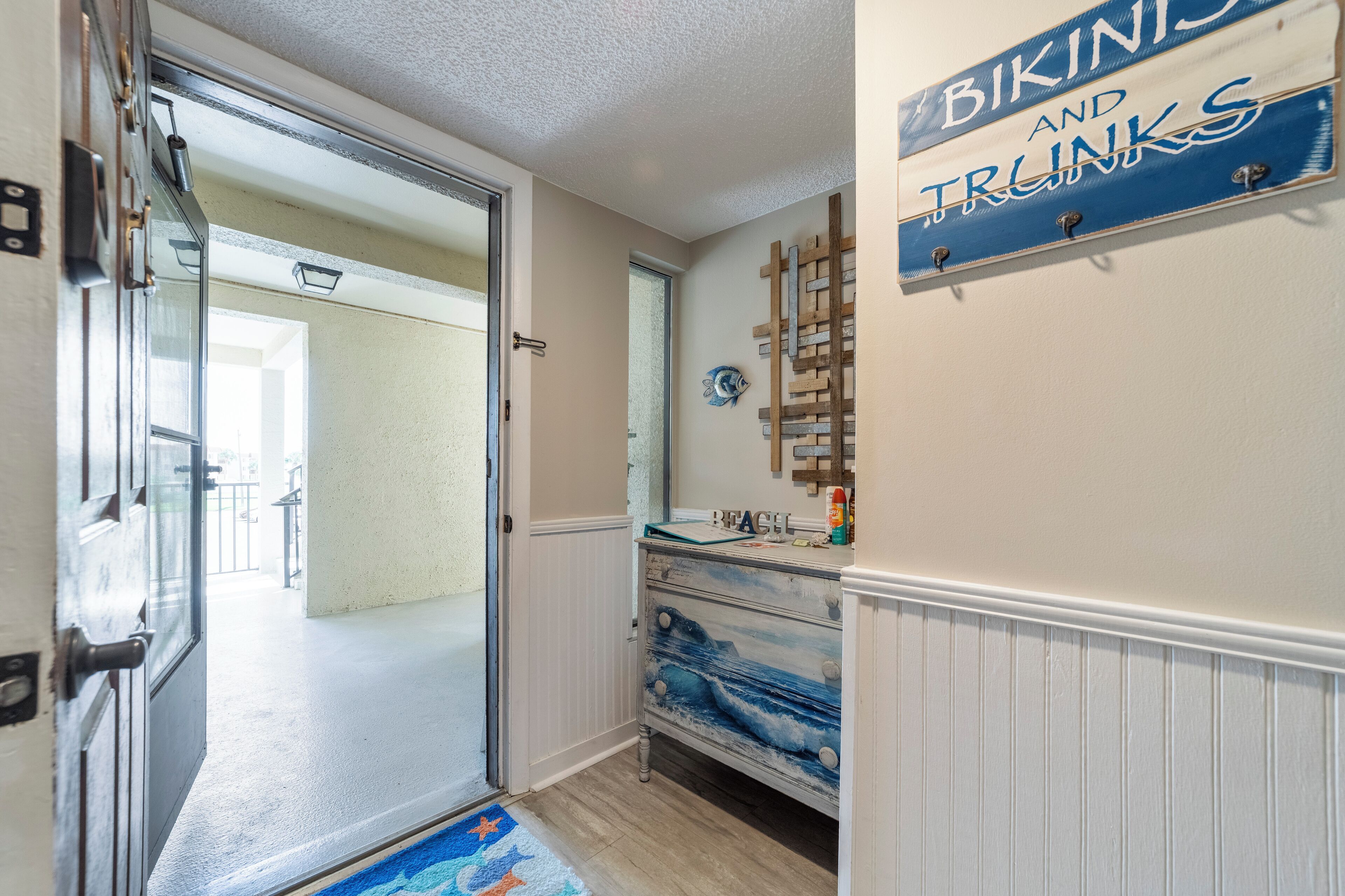 A hallway with a door leading to a room, a wall-mounted sign reading "Bikinis and Trunks", and a wooden rack with items on it.