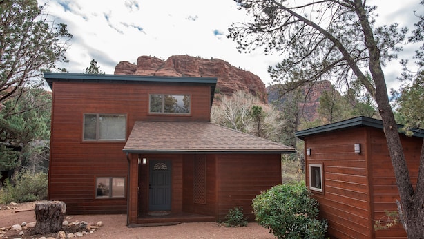 A two-story wooden cabin with a front porch, a small attached shed, and a mountainous backdrop.