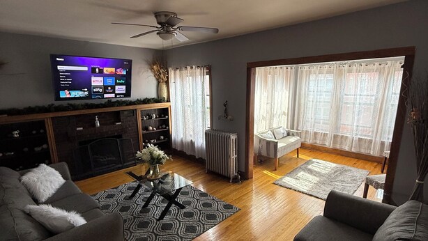 A living room with a flat-screen TV, grey sofas, a coffee table, a fireplace, and a large window with curtains. - Hyde Park/ Bronzeville Apartment 3E (Chicago)