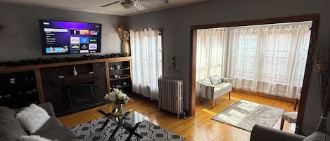 Una sala de estar con televisor de pantalla plana, sofás grises, una mesa de centro, chimenea y un ventanal con cortinas.