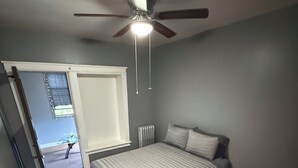 A bedroom with a bed, a ceiling fan, a radiator, a white rug, and a door leading to an outdoor area. - Hyde Park/ Bronzeville Apartment 3E (Chicago)