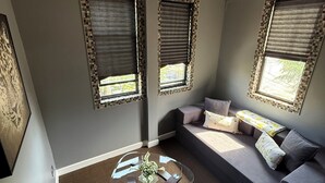 A modern living room with a glass coffee table, a grey sofa, and two windows with patterned blinds. - Hyde Park/ Bronzeville Apartment 3E (Chicago)