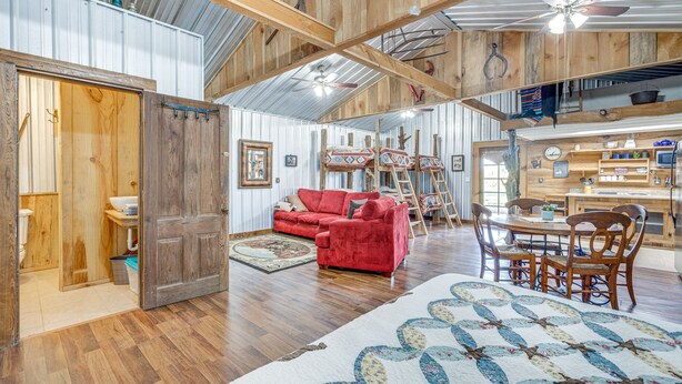 A spacious living area with a red sofa, wooden dining table, and chairs. A large quilt is on the floor, and there is a wooden door leading to another room.