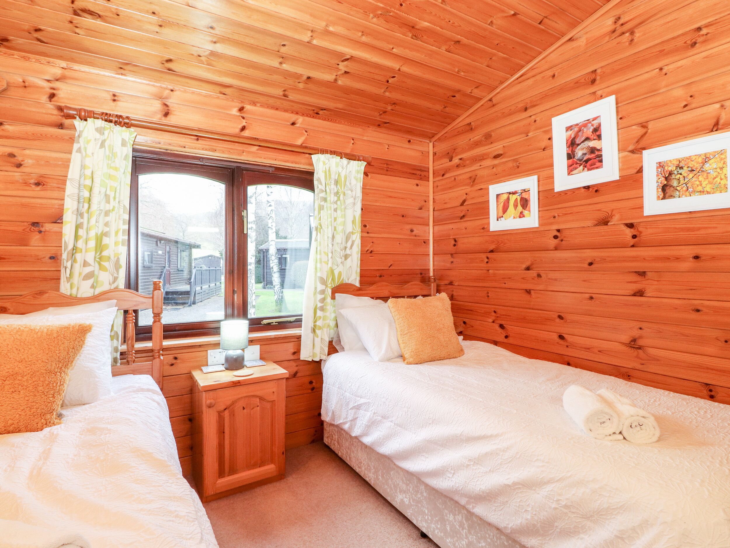 Ein Schlafzimmer einer Holzhütte mit zwei Betten, einem Nachttisch, einem Fenster mit Vorhängen und gerahmten Bildern an der Wand.
