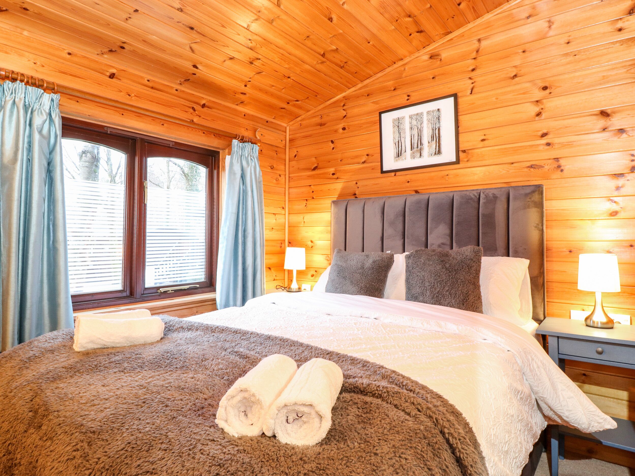 A wooden cabin bedroom with a bed, two bedside lamps, a framed picture on the wall, and a window with curtains.