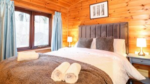 A wooden cabin bedroom with a bed, two bedside lamps, a framed picture on the wall, and a window with curtains. - Latrigg Lodge (Keswick)