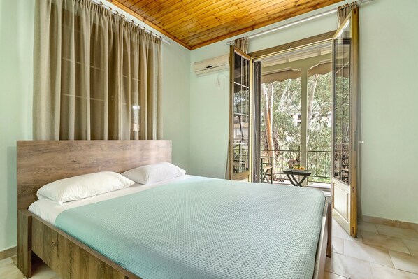 Quarto com cabeceira de madeira, cama com travesseiros brancos, colcha verde-água e varanda com mesa e cadeiras.