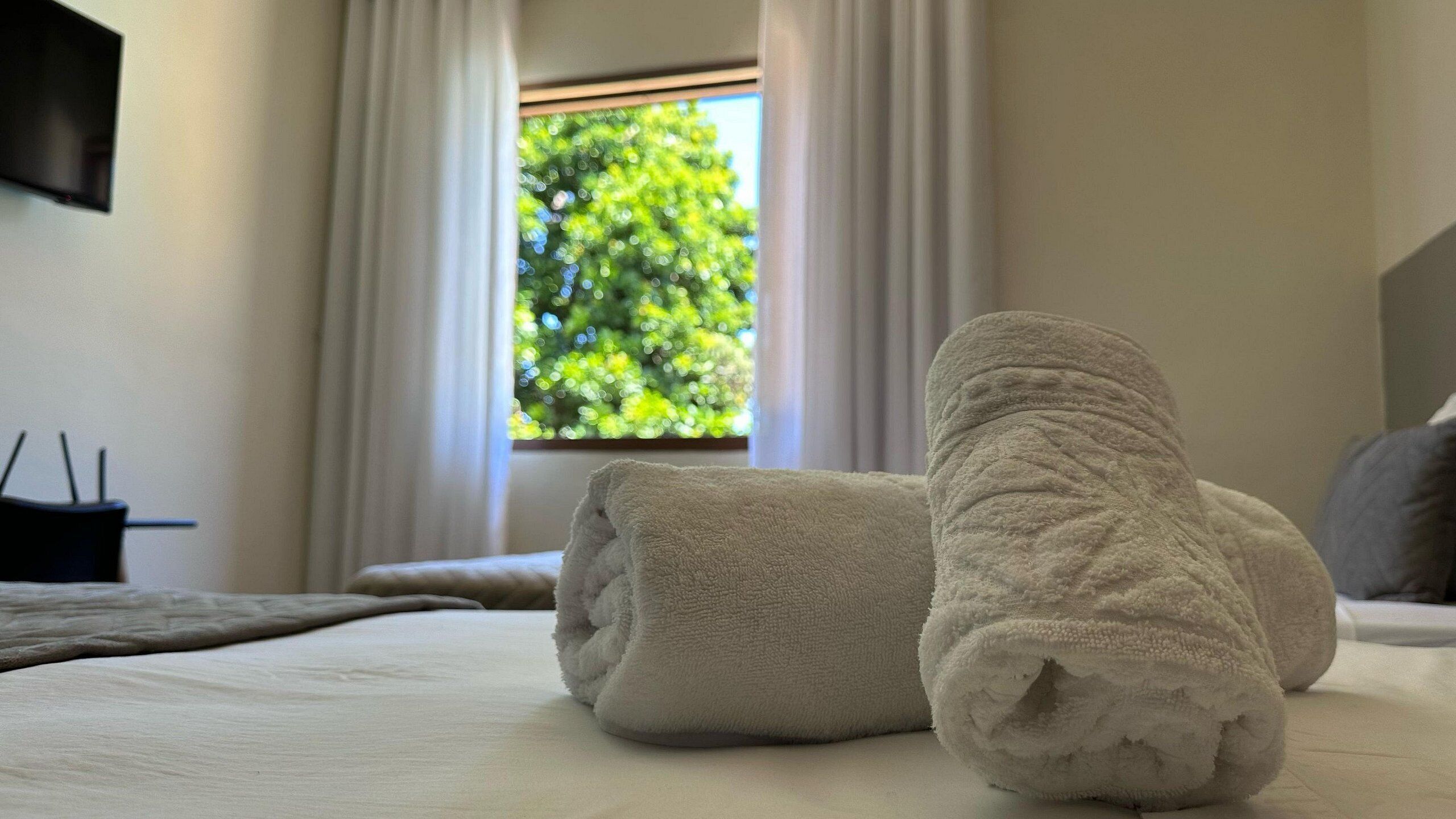 A neatly made bed with rolled towels and a window with a view of greenery.