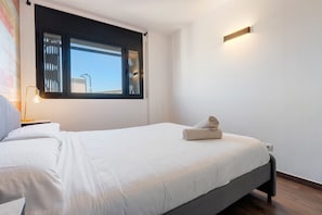 Un lit simple avec une couette blanche, une lampe de chevet et une fenêtre avec des stores.