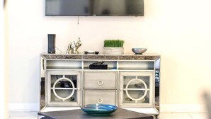 A modern living room with a flat-screen TV mounted on the wall, a marble-patterned cabinet with glass doors, and a decorative bowl on the table. - Family Townhome near Disney! Patio w/Pvt Splash Pool, Modern Decor,Free wifi, Resort w/waterpark! (Kissimmee)