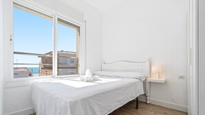 Un dormitorio con cama, una mesita de noche con lámpara, ventana con vistas a edificios y suelo de madera.