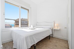 Un dormitorio con cama, una mesita de noche con lámpara, ventana con vistas a edificios y suelo de madera.