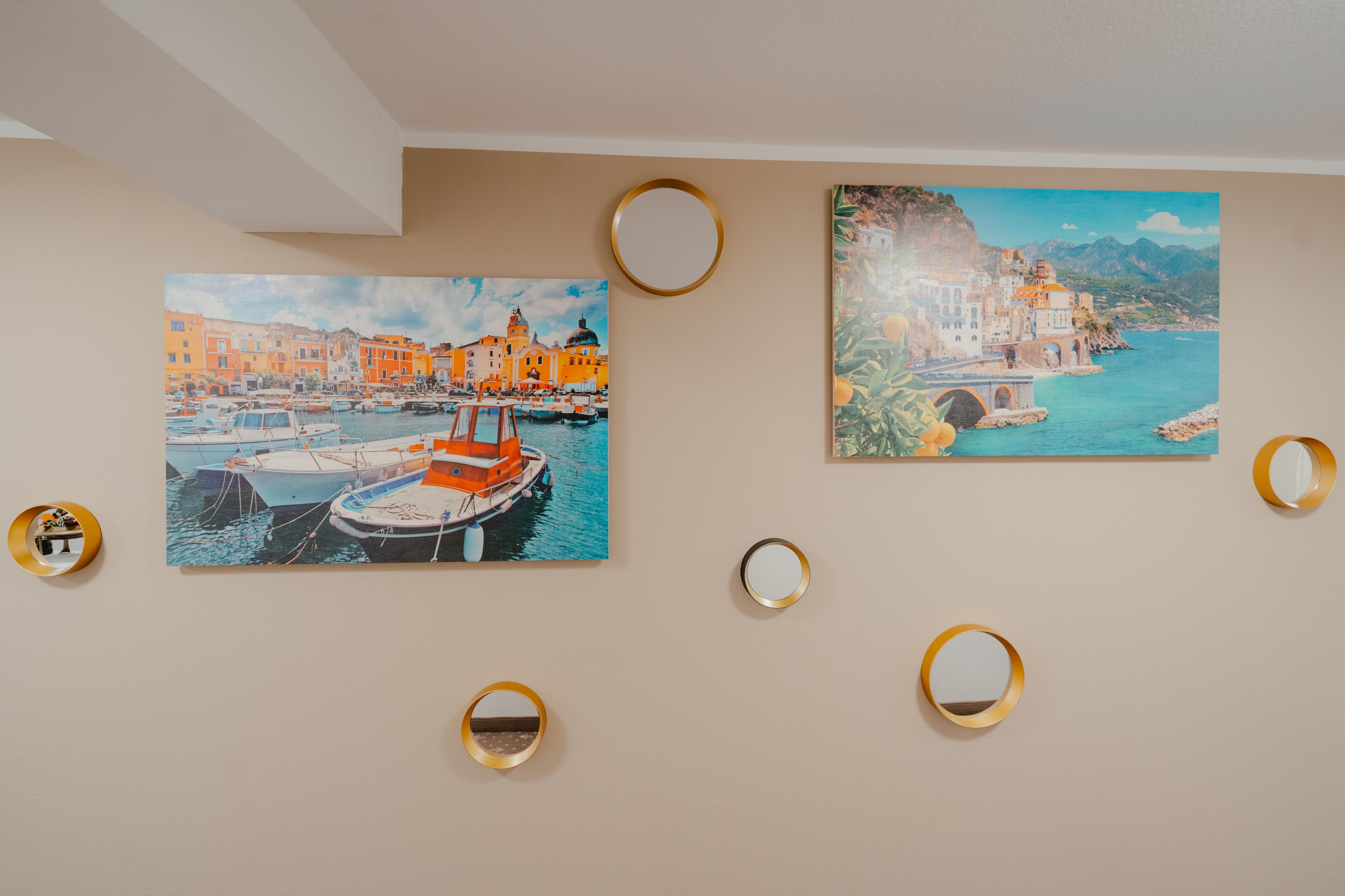 Two paintings of coastal scenes with boats, framed by gold rings on a plain wall.