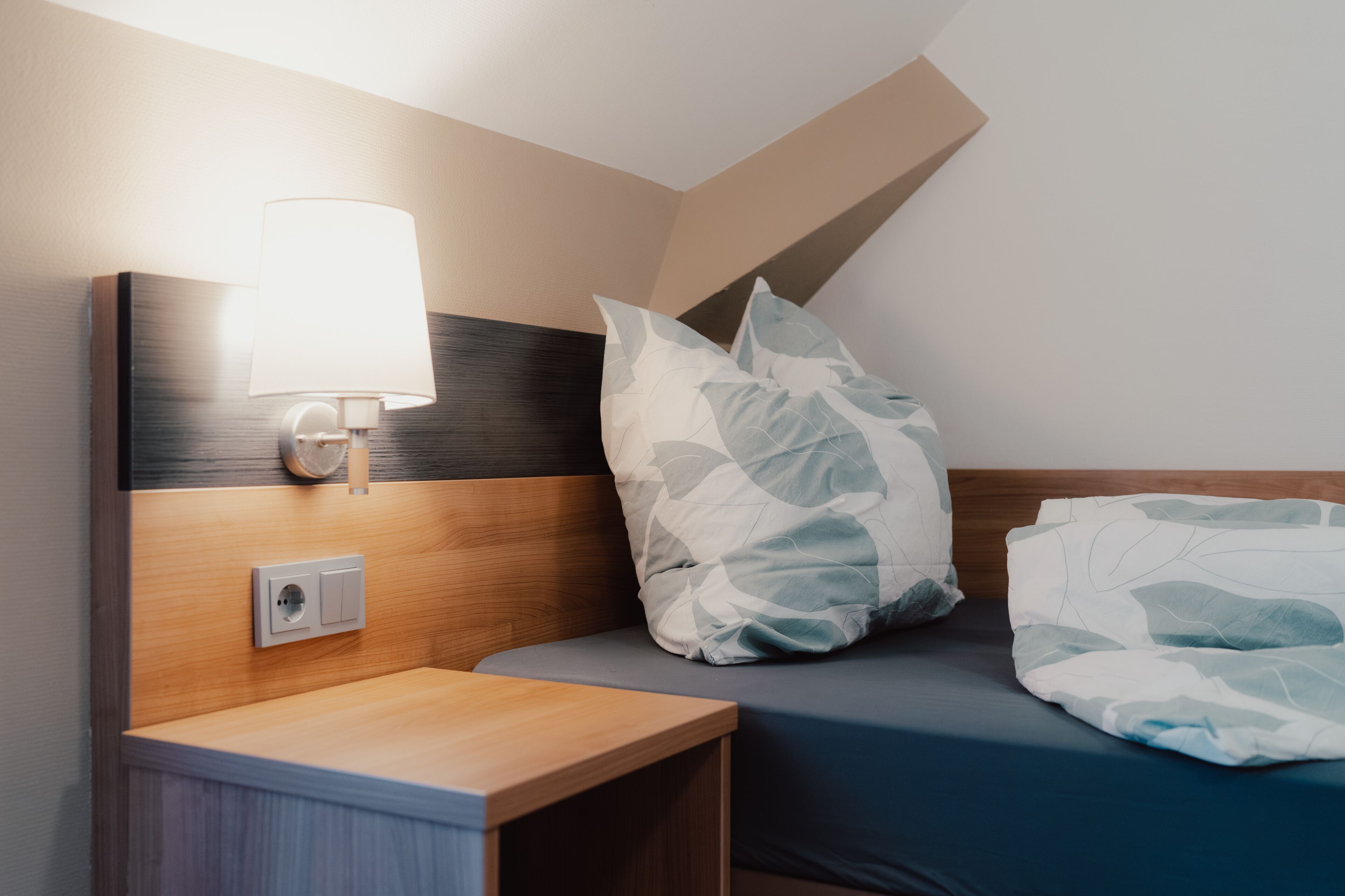A neatly made bed with a patterned pillow, a bedside lamp, and a power outlet on a wooden nightstand.