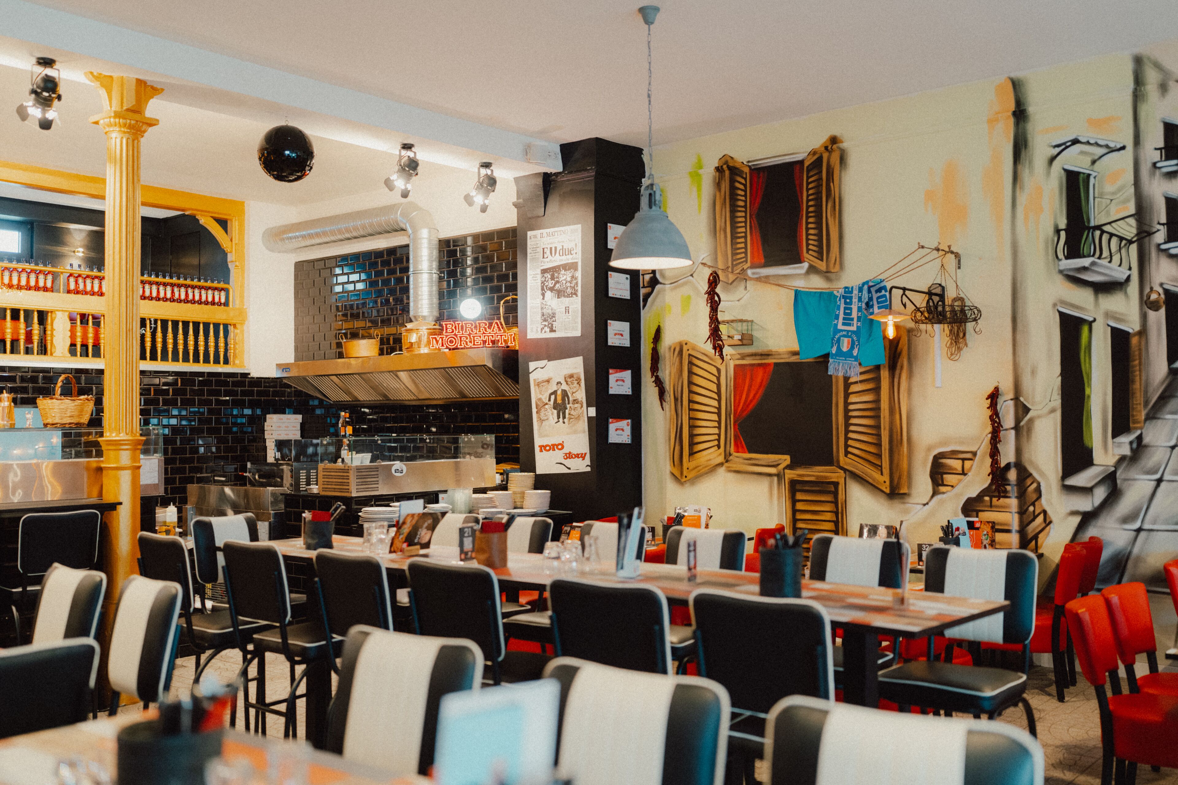 A restaurant interior with multiple dining tables and chairs, a kitchen area, and decorative elements like a column and wall art.