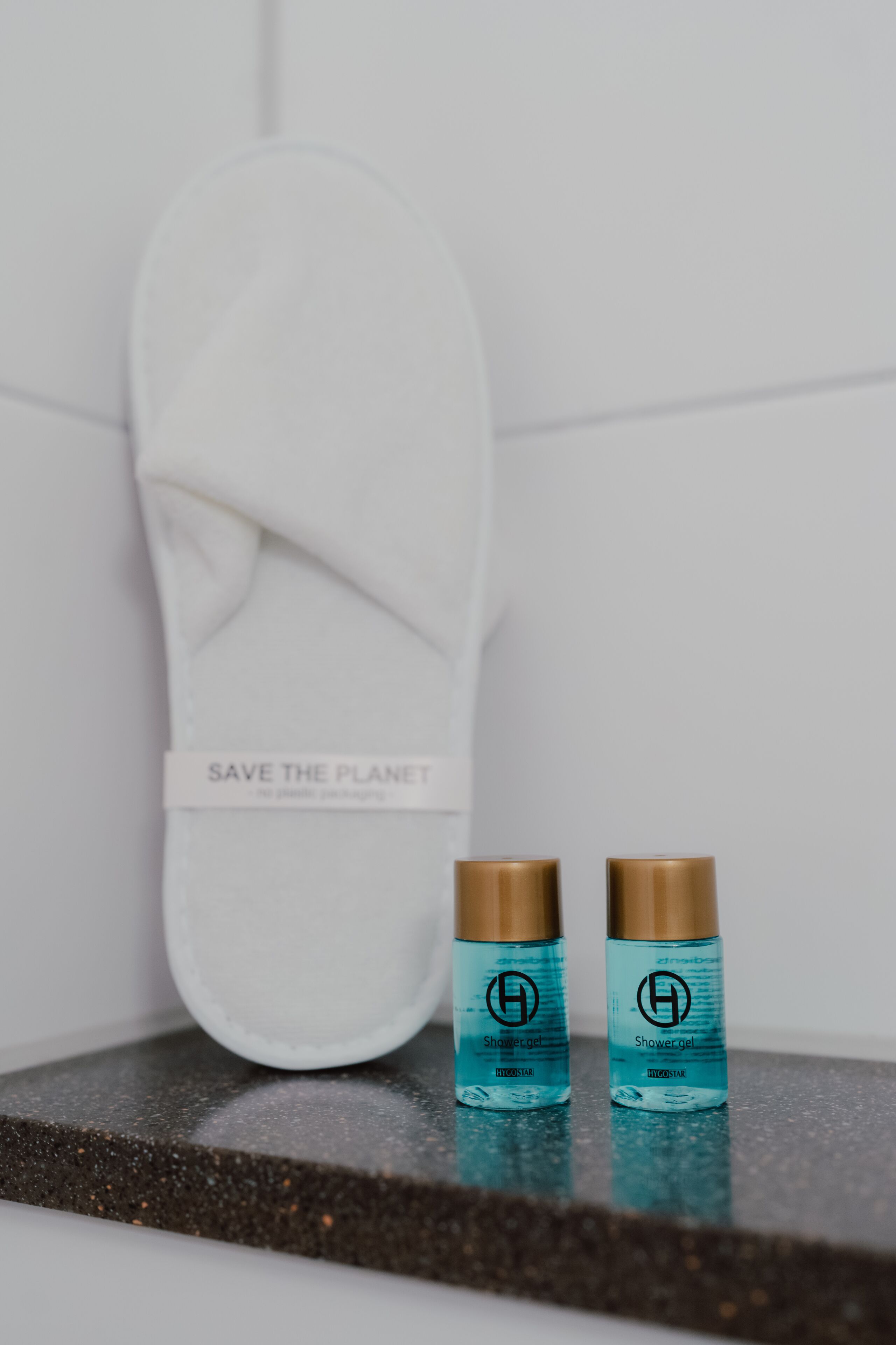 Un baño con una toalla blanca colgada en un perchero, dos botellas de gel de ducha y una etiqueta que dice "SAVE THE PLANET" en la toalla.