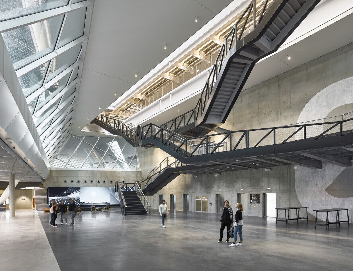 Een ruim interieur met een hoge plafond, meerdere niveaus verbonden door trappen, en een groot digitaal scherm.