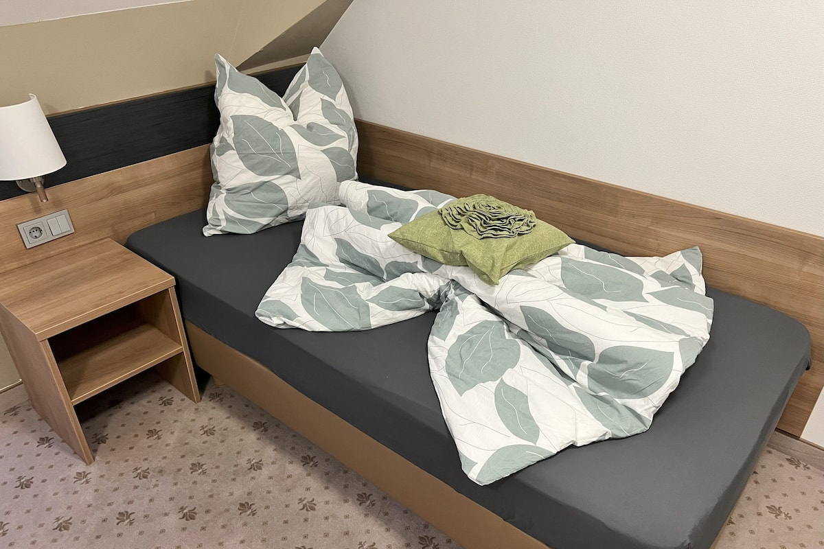 Un lit avec un matelas gris, des oreillers à motifs de feuilles, un plaid vert et une table de chevet avec une lampe.