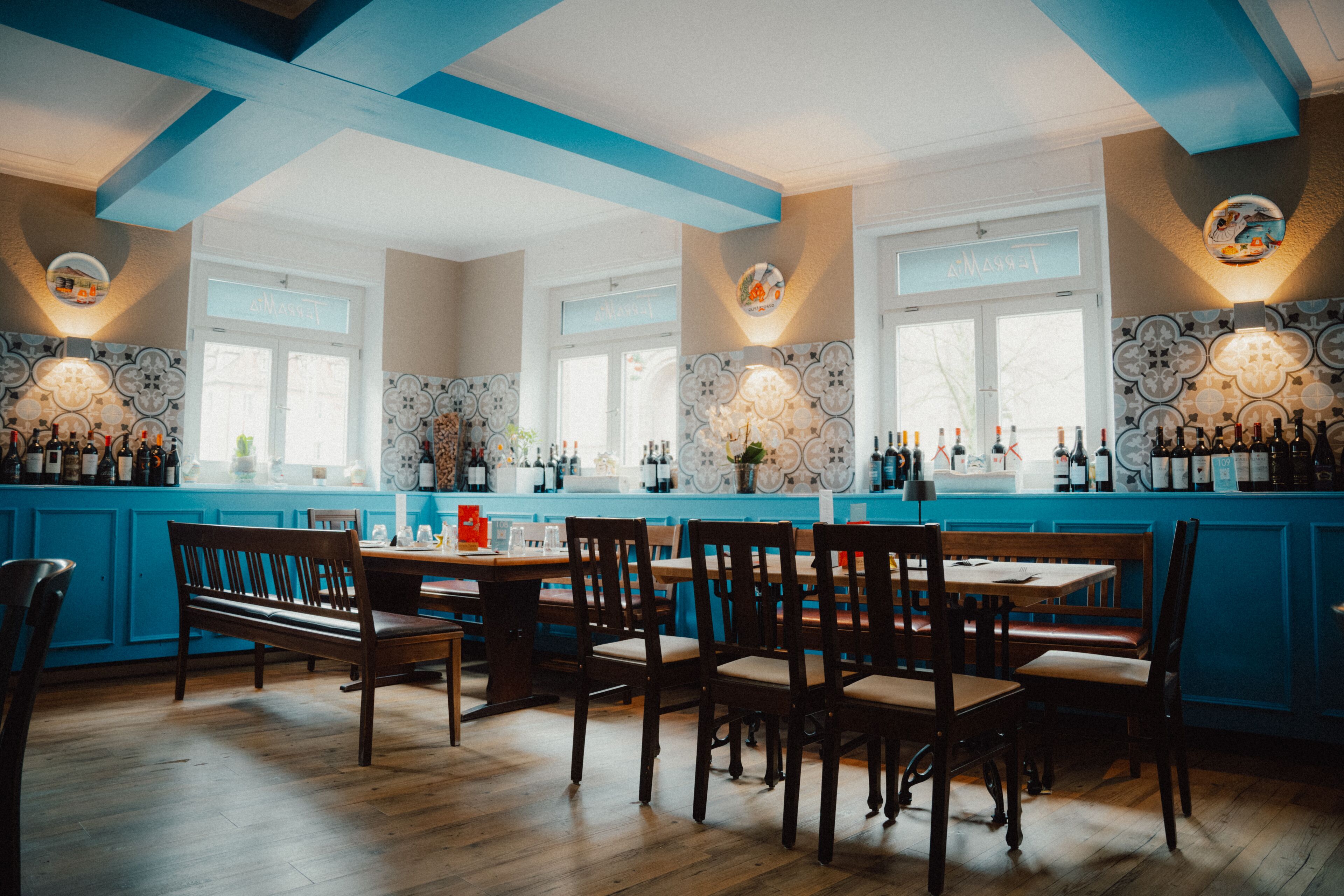 A restaurant interior with wooden tables and chairs, a blue bar counter, and decorative wall tiles.