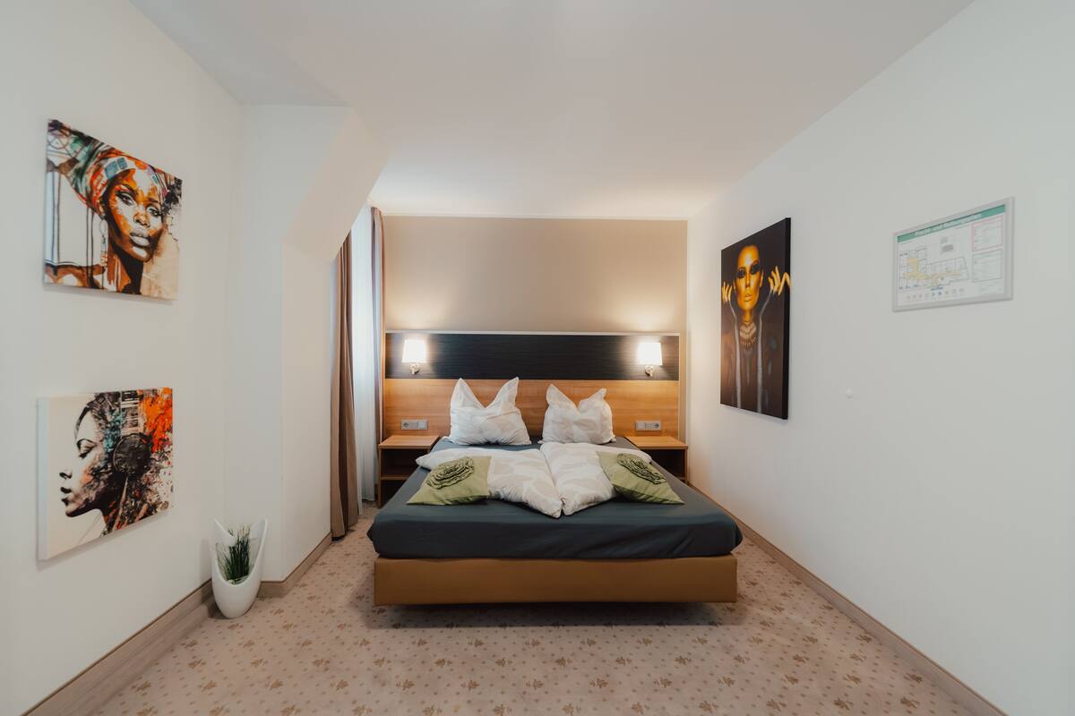 Une chambre d’hôtel moderne avec un grand lit, une tête de lit dotée d’un éclairage intégré, une petite plante en pot et des œuvres d’art accrochées aux murs.