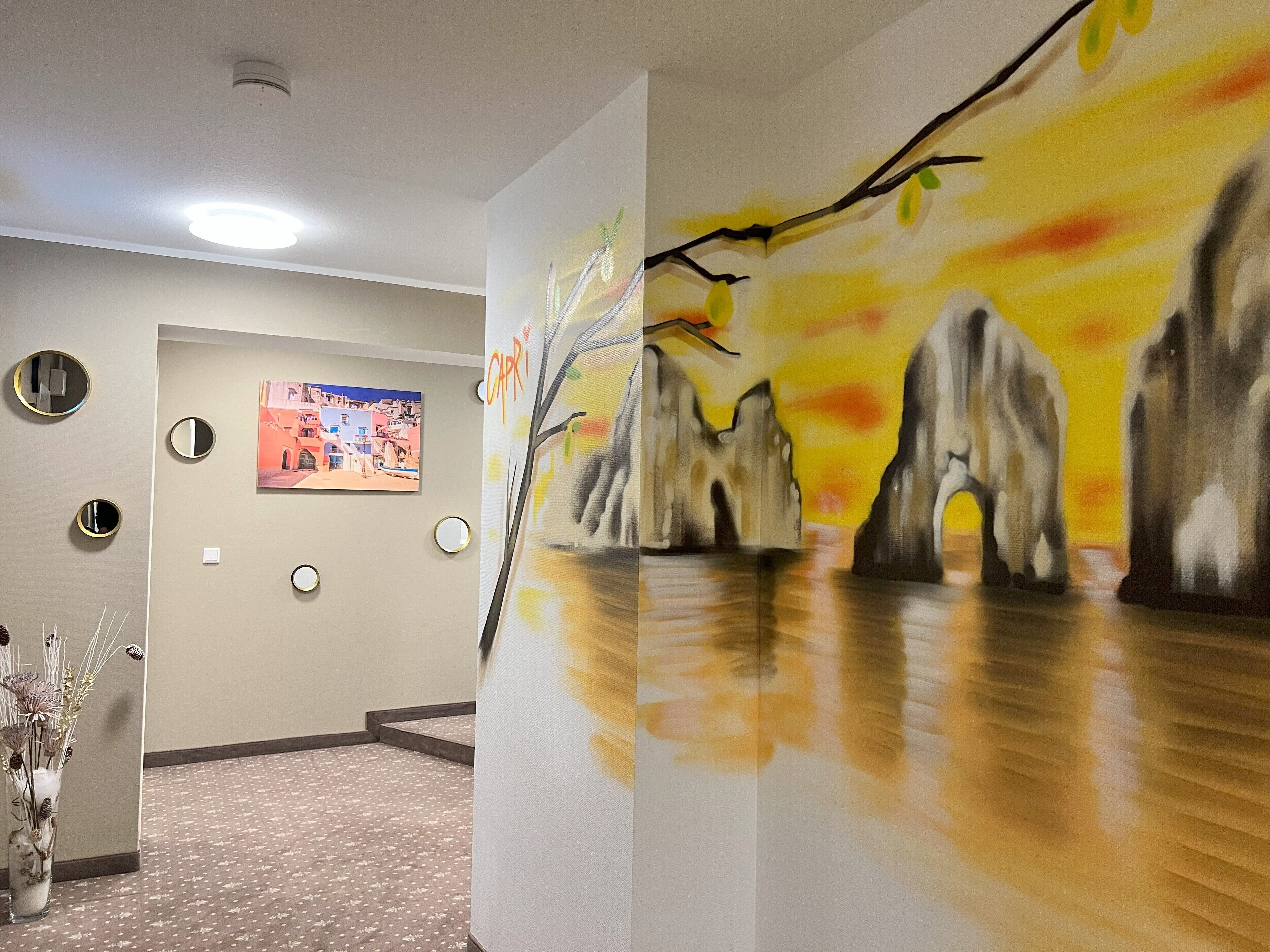 A hallway with a painting of a tree branch, a framed photograph, and a vase with dried flowers.