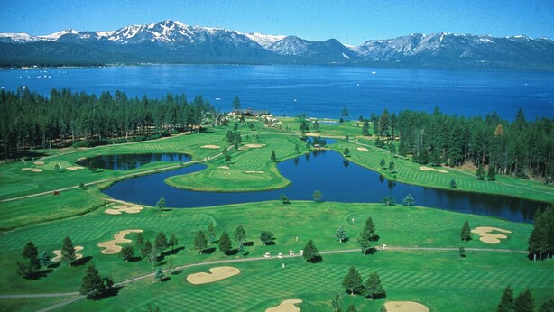 Un campo de golf con varios estanques, rodeado de bosques densos y montañas al fondo.