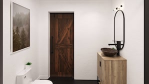 Un baño moderno con inodoro blanco, puerta de madera, mueble de madera con lavabo integrado y un cuadro enmarcado en la pared.
