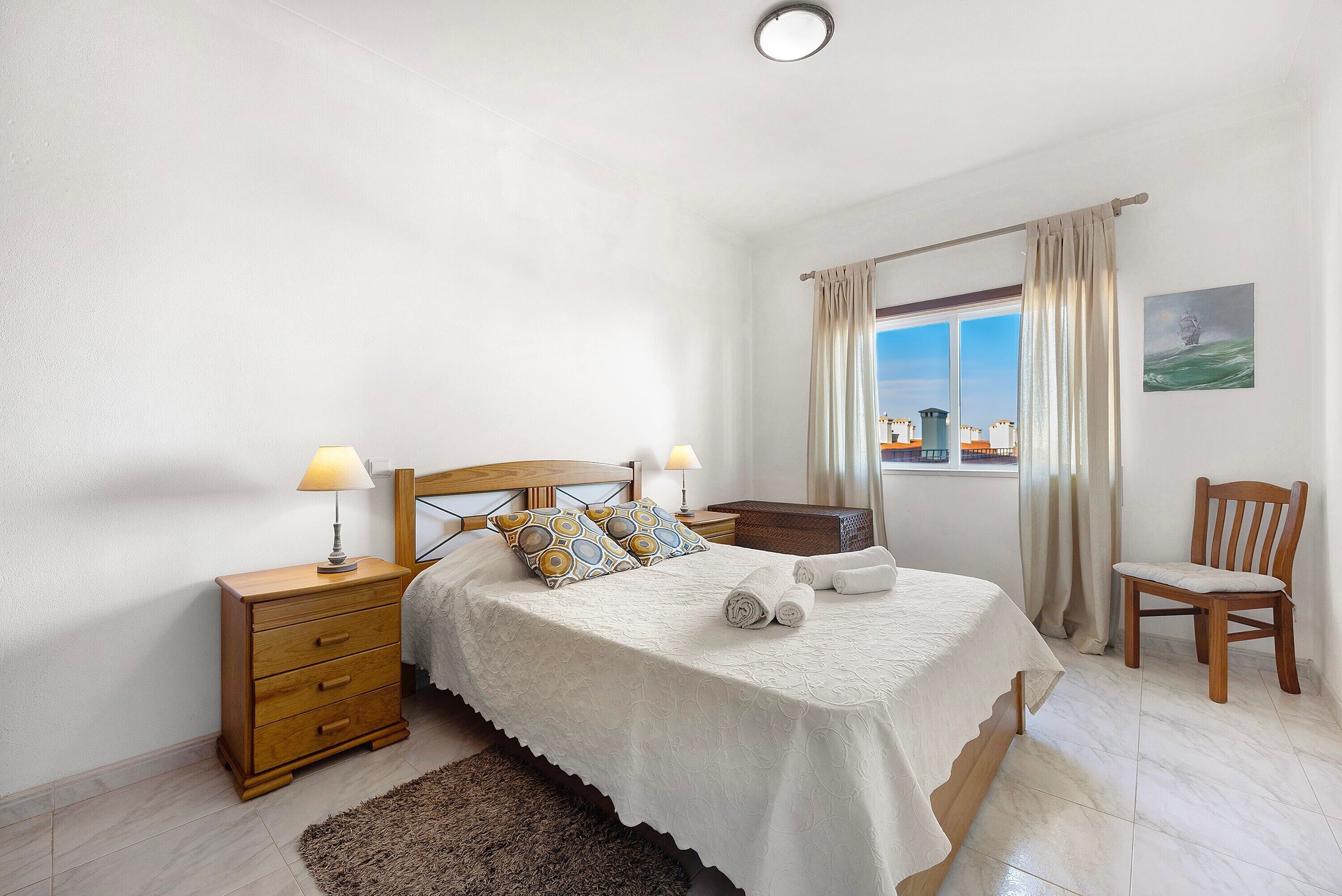 A bedroom with a bed, two bedside tables, a chair, a window with curtains, and a painting on the wall.