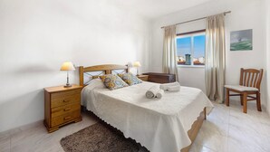 Un dormitorio con cama, dos mesitas de noche, una silla, una ventana con cortinas y un cuadro en la pared.