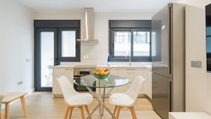 Une cuisine moderne avec une table à manger en verre, deux chaises blanches, un réfrigérateur en acier inoxydable et une fenêtre donnant sur la ville.
