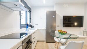 Une cuisine moderne avec des meubles blancs, un réfrigérateur en acier inoxydable, une table à manger en verre accompagnée de deux chaises blanches, et un téléviseur à écran plat fixé au mur.
