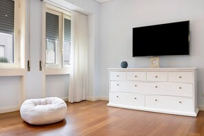 Une pièce avec une commode blanche, un téléviseur à écran plat, un fauteuil en forme de poire et une fenêtre avec des stores.