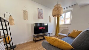 A living room with a flat-screen TV, a grey sofa with yellow cushions, a black metal shelf, and a macramé wall hanging. - Top Moderne Wohnung im Stadtkern von Oettingen (Oettingen in Bayern)