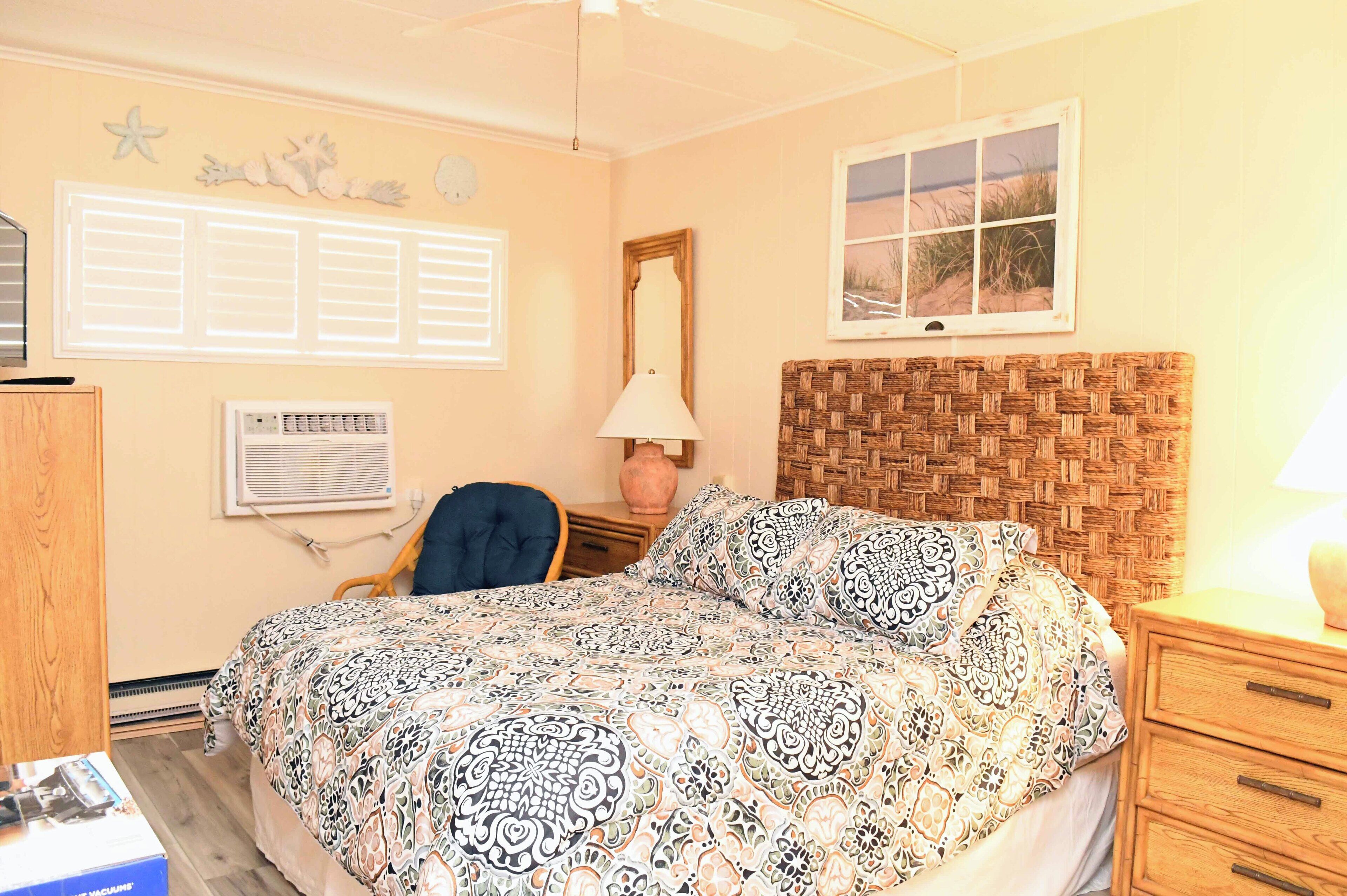 A bedroom with a patterned bedspread, a woven headboard, a nightstand with a lamp, a chair, a wall clock, and a window with a beach view.