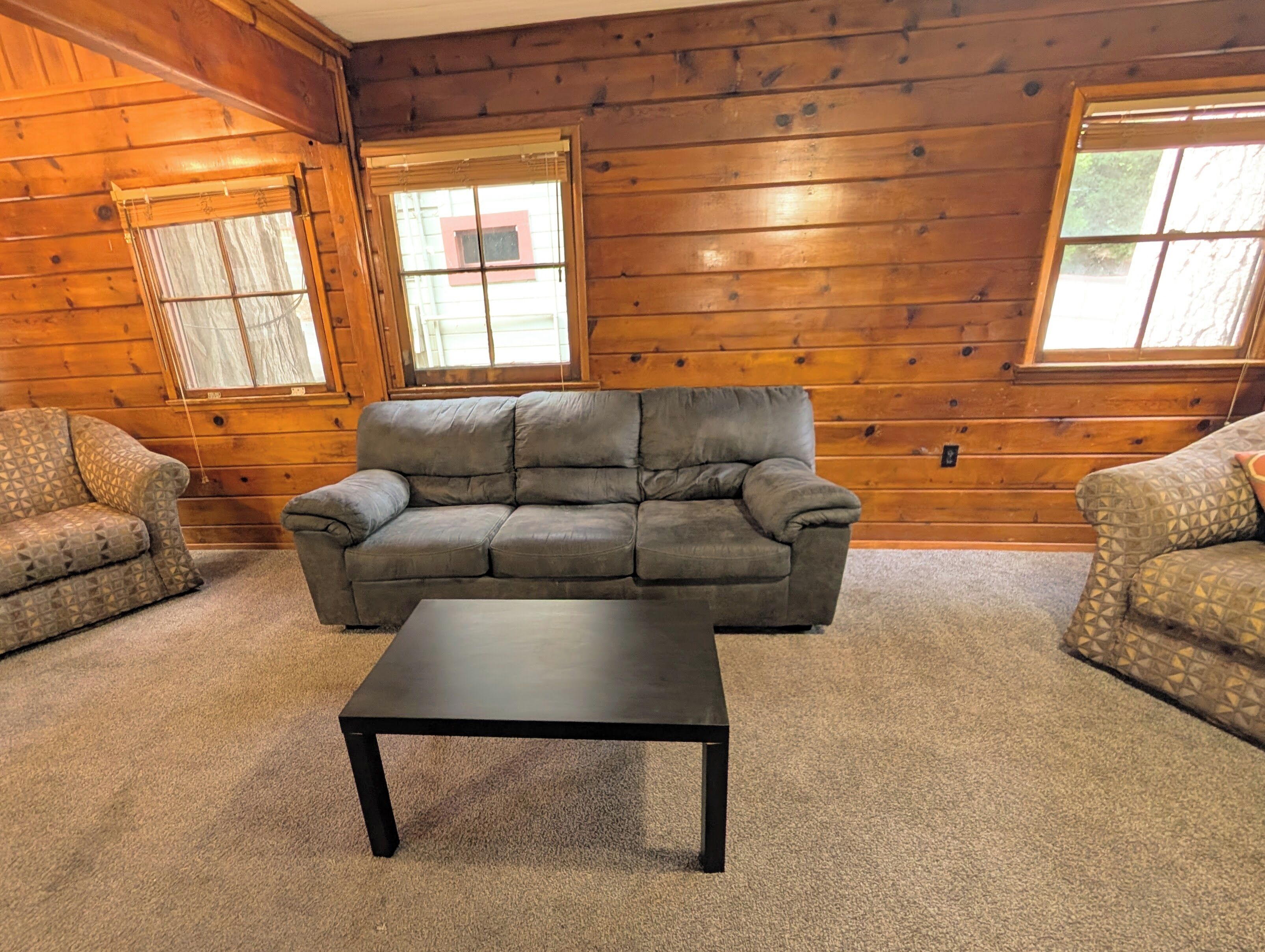 Ein Zimmer mit grauem Sofa, zwei gemusterten Sessel, einem schwarzen Couchtisch und Holzwänden mit Fenstern.